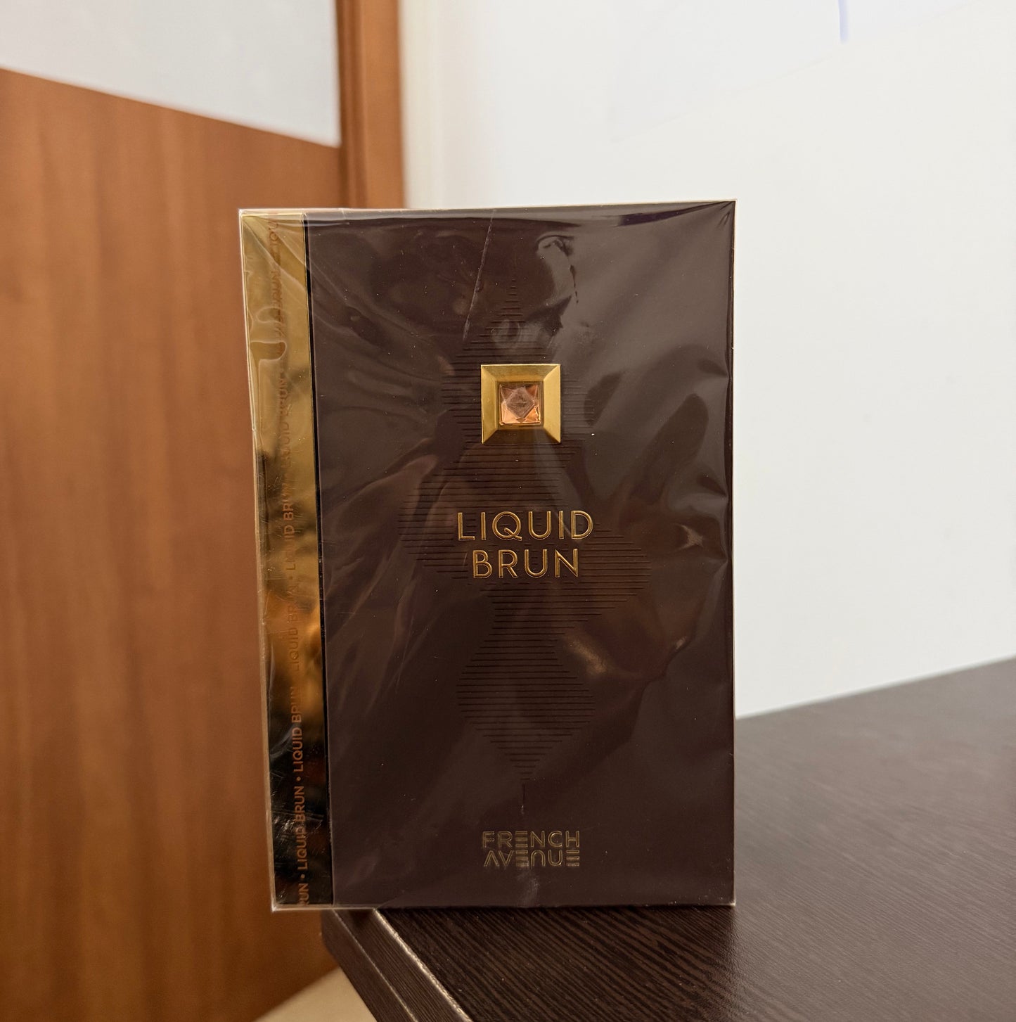 A boxed fragrance product labeled 'LIQUID BRUN' and 'FRESH' in gold lettering on a dark brown box, photographed indoors against a white wall and wood floor.