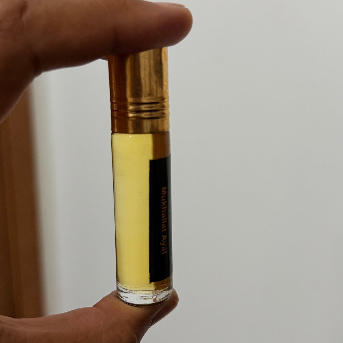 A hand holding a cylindrical glass vial containing pale yellow powder or liquid, with a gold cap and a black label that reads 'Mukhasth Ayal'.