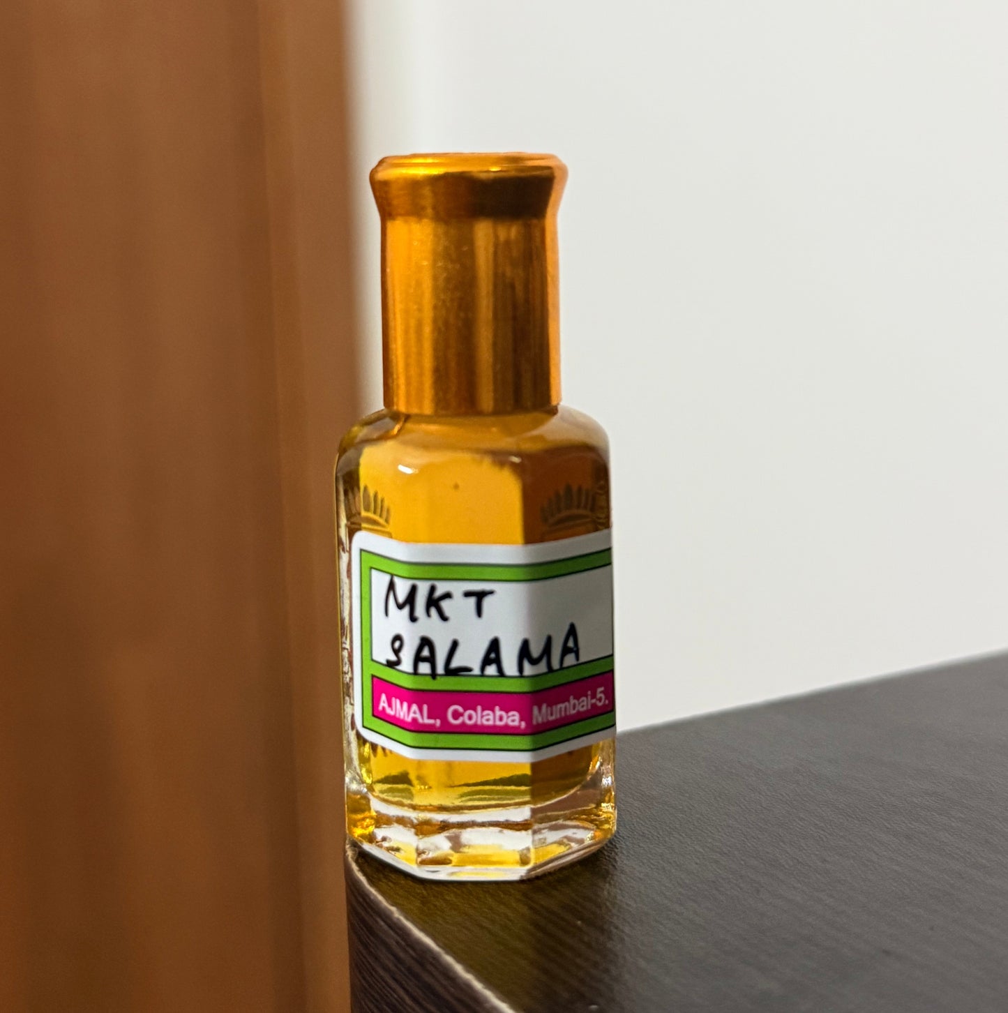 A small clear glass bottle filled with amber liquid, with a gold-toned cap, and a white label reading 'MKT SALAMA', resting on a wooden surface against a dark gray background.