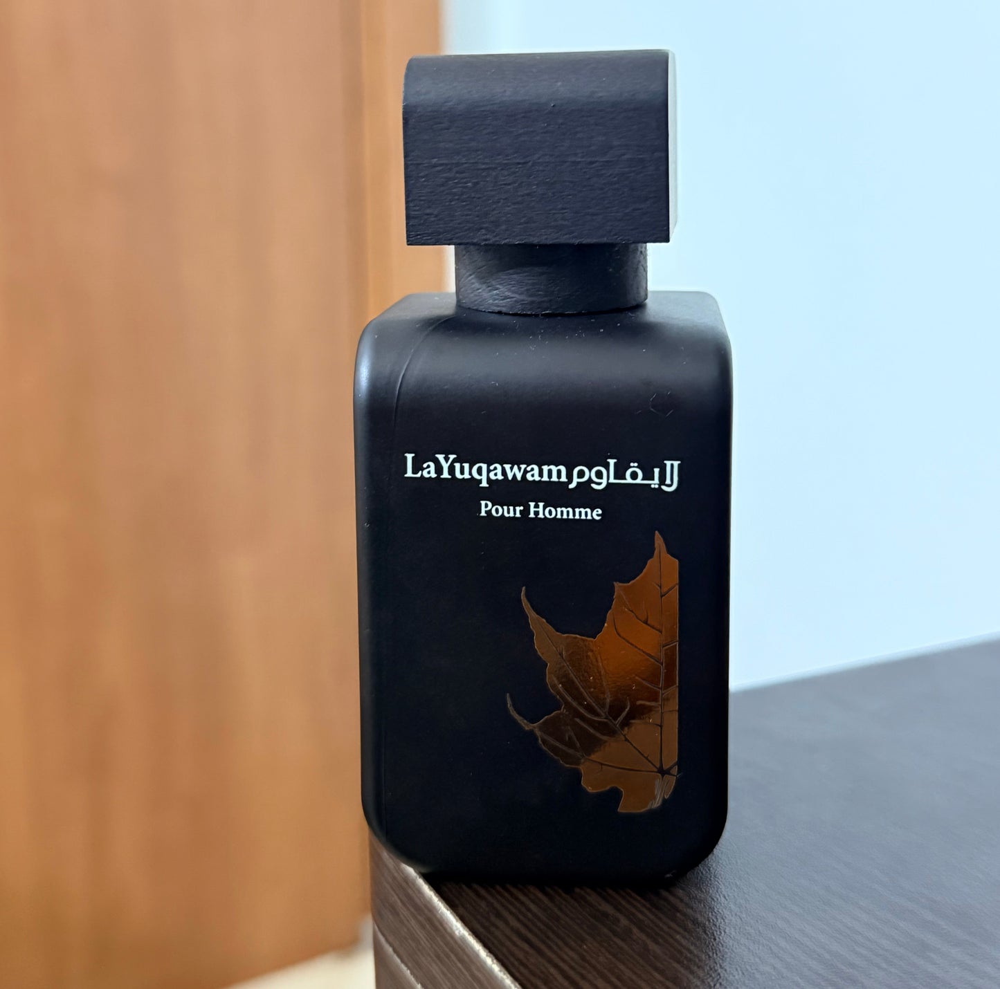 A black rectangular perfume bottle labeled 'LaYuqawam Pour Homme' with a gold leaf graphic, standing on a dark wooden surface against a light blue wall and wood panel background.