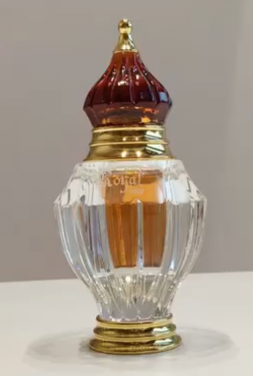 A vintage-style perfume bottle with a faceted clear glass body, a gold-toned base and neck, and a dark reddish-brown pointed cap, shown on a light surface against a neutral background.