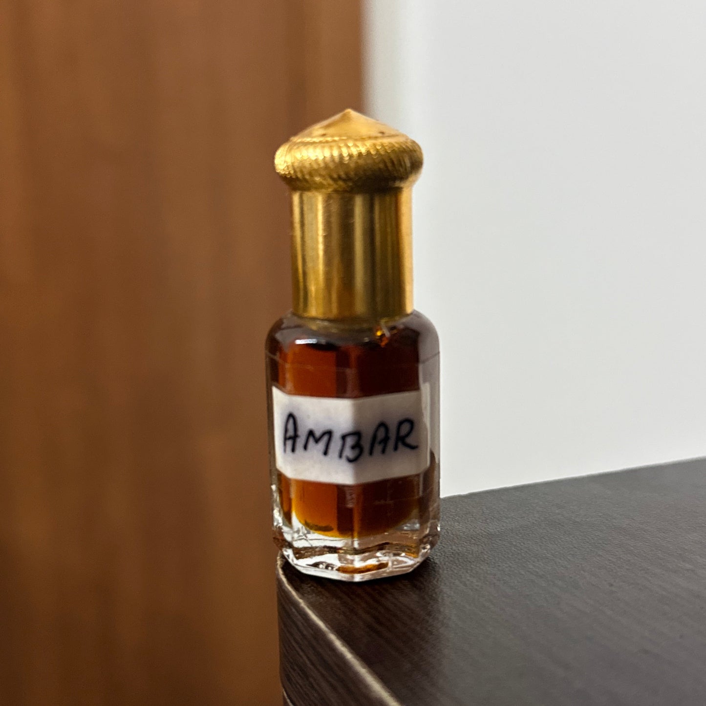 A small glass perfume bottle with a gold cap and a handwritten white label reading 'AMBAR', resting on a dark wooden surface against a plain wall.