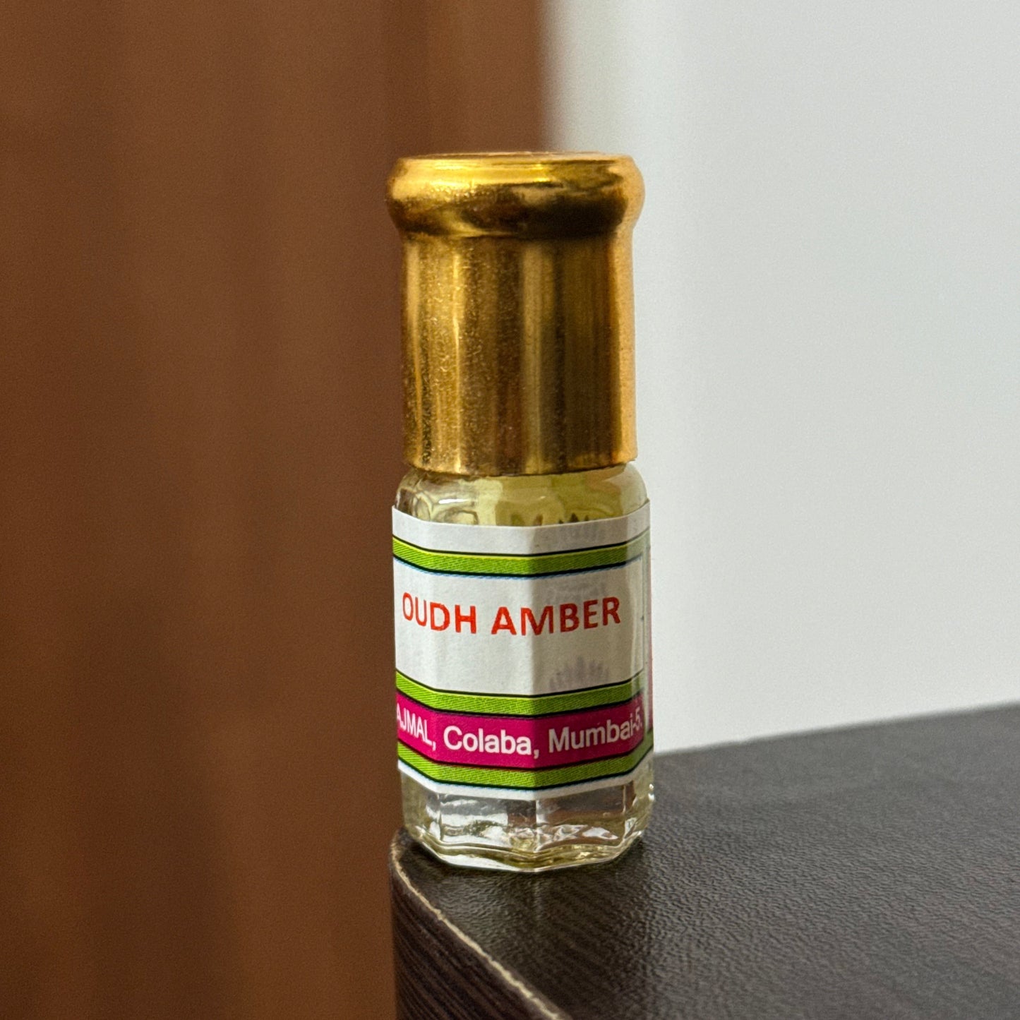 A small glass bottle of Oudh Amber oil with a gold-colored cap, resting on a dark textured surface against a neutral background.