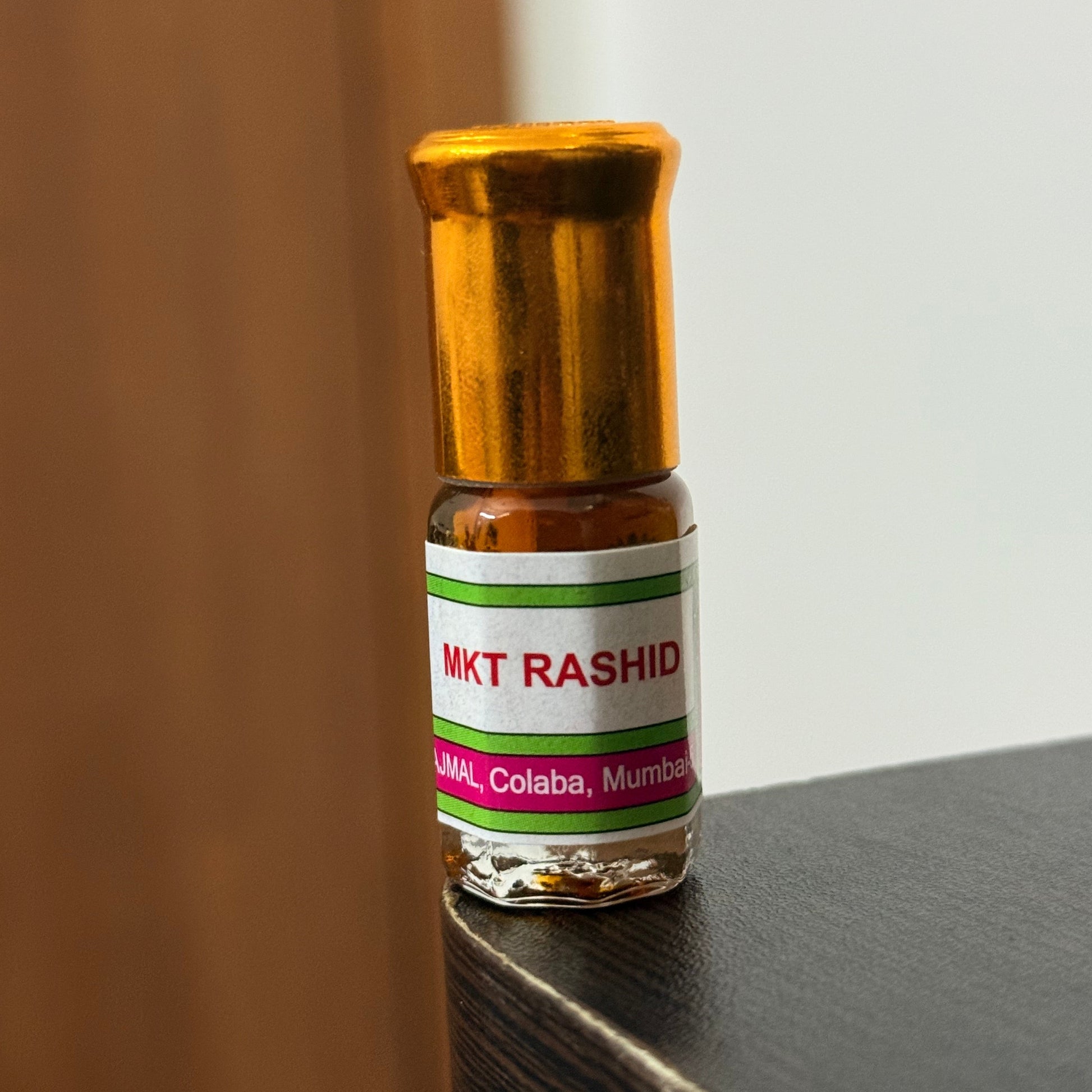 A small glass bottle of MKT Rashid hair growth serum with a gold cap, resting on a dark textured surface against a light background.