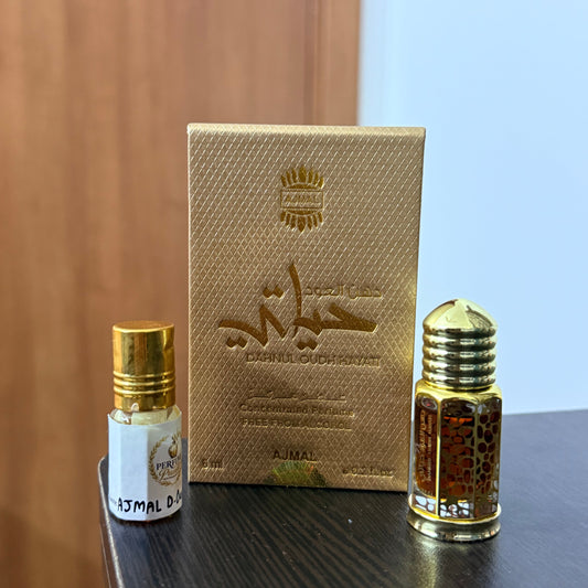 A small gold rectangular box with embossed script, shown beside two cylindrical bottles with gold caps and clear bodies containing a golden liquid, photographed on a dark tabletop with a light wall in the background.