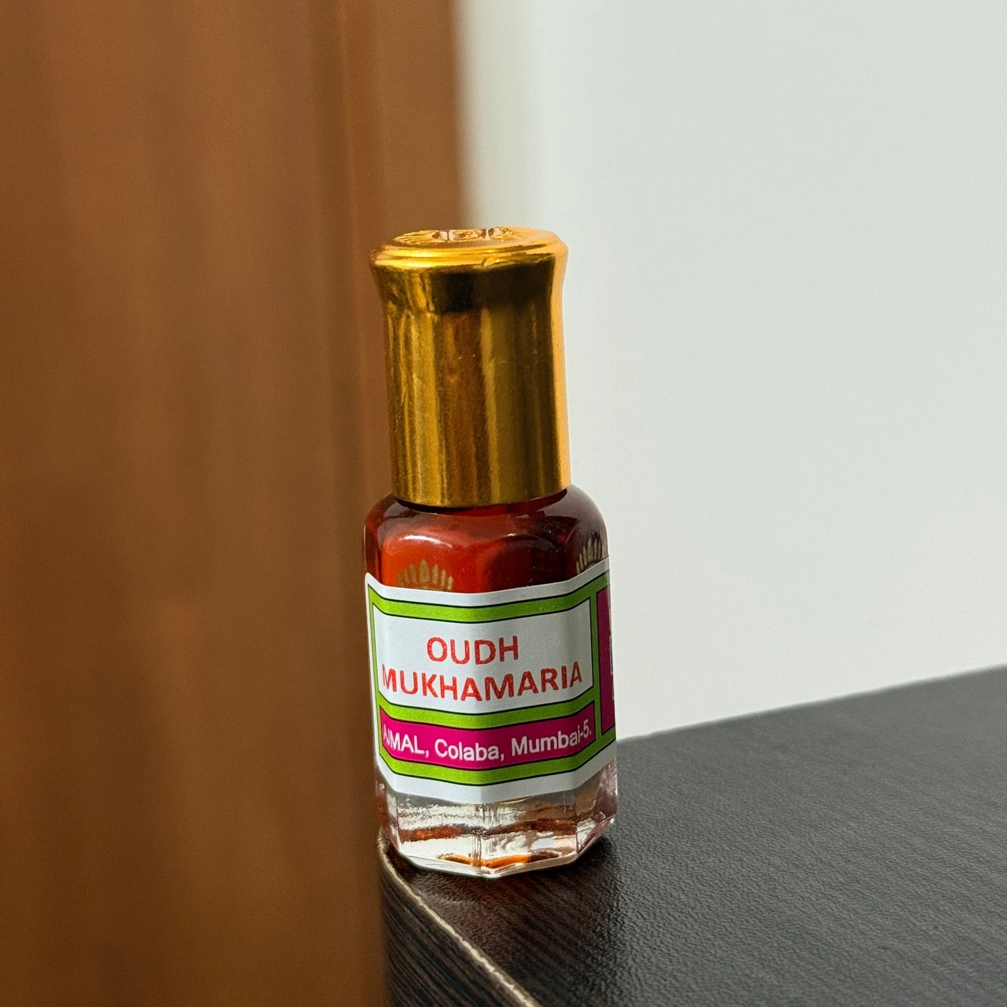 A small bottle of Oudh Mukhamri herbal oil with a gold cap and a white label, resting on a wooden surface against a dark gray background.