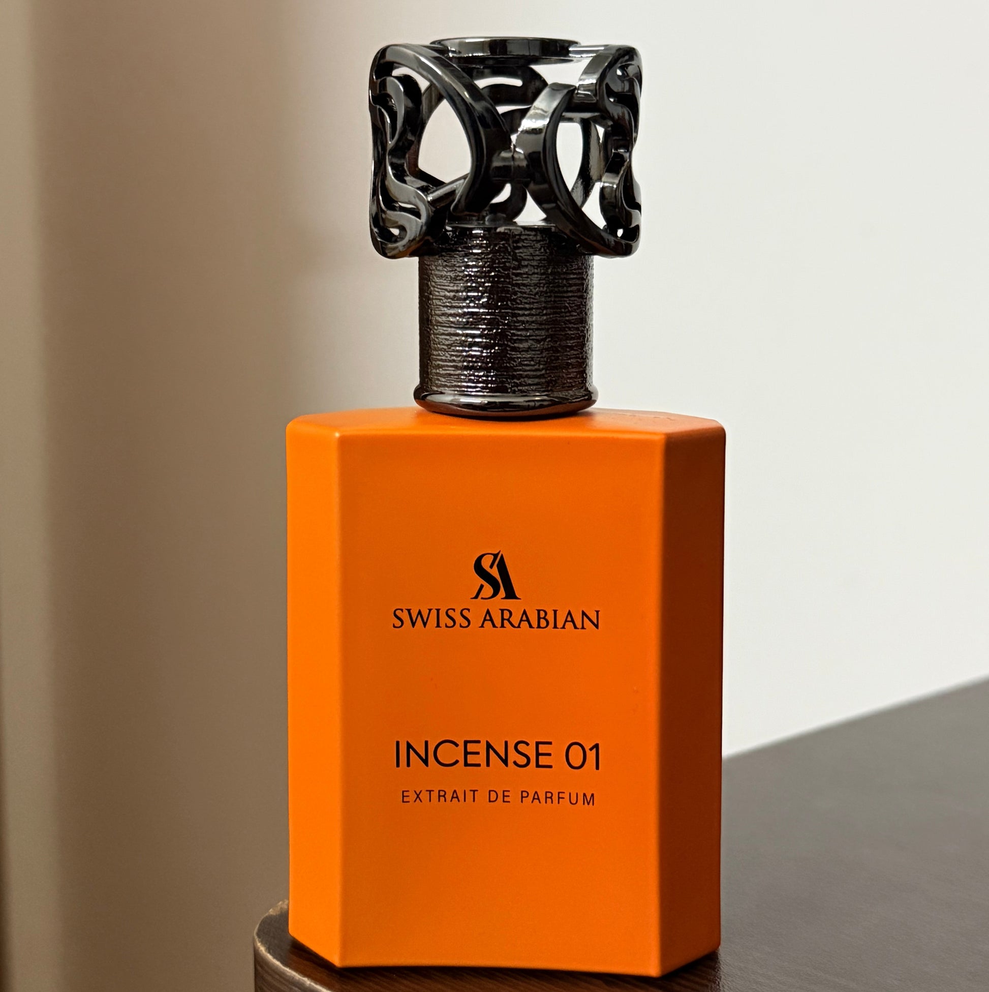 Swiss Arabian Incense 01 perfume bottle in vibrant orange with a black ornate cap, displayed on a dark wooden surface against a neutral beige background.