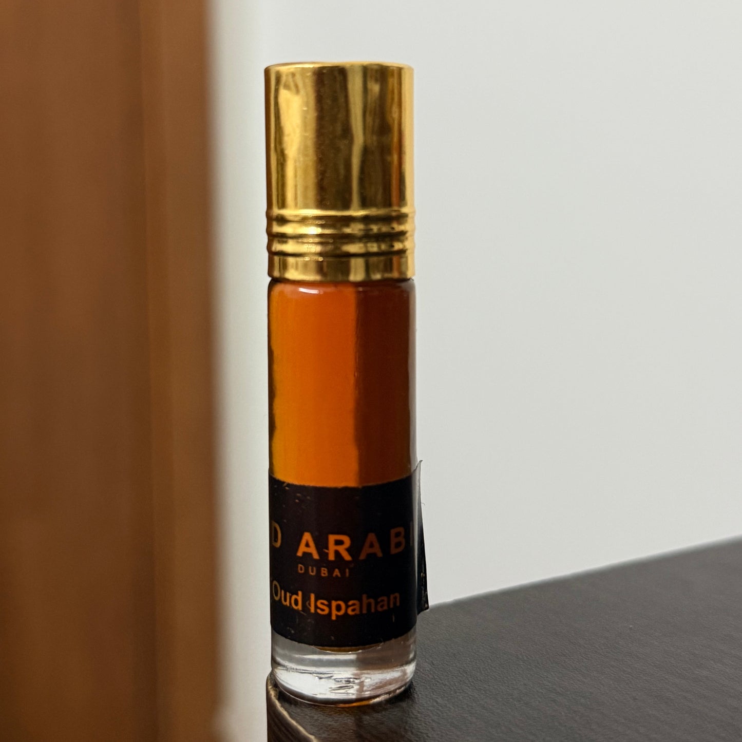 A small cylindrical glass bottle of Arabic oud Isfahan perfume oil with a shiny gold cap and a black label, standing on a dark wooden surface against a neutral background.