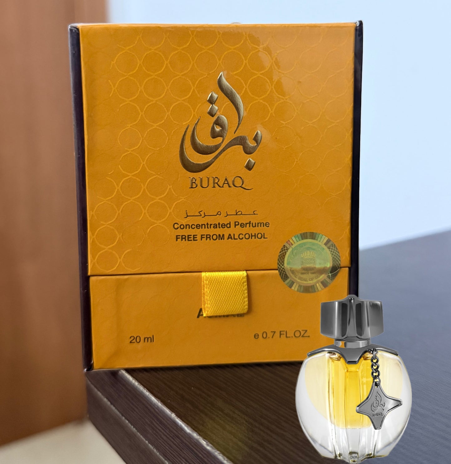 Buraq concentrated perfume box in golden yellow packaging next to a small clear glass perfume bottle with yellow liquid and a silver cap, on a dark wooden surface.