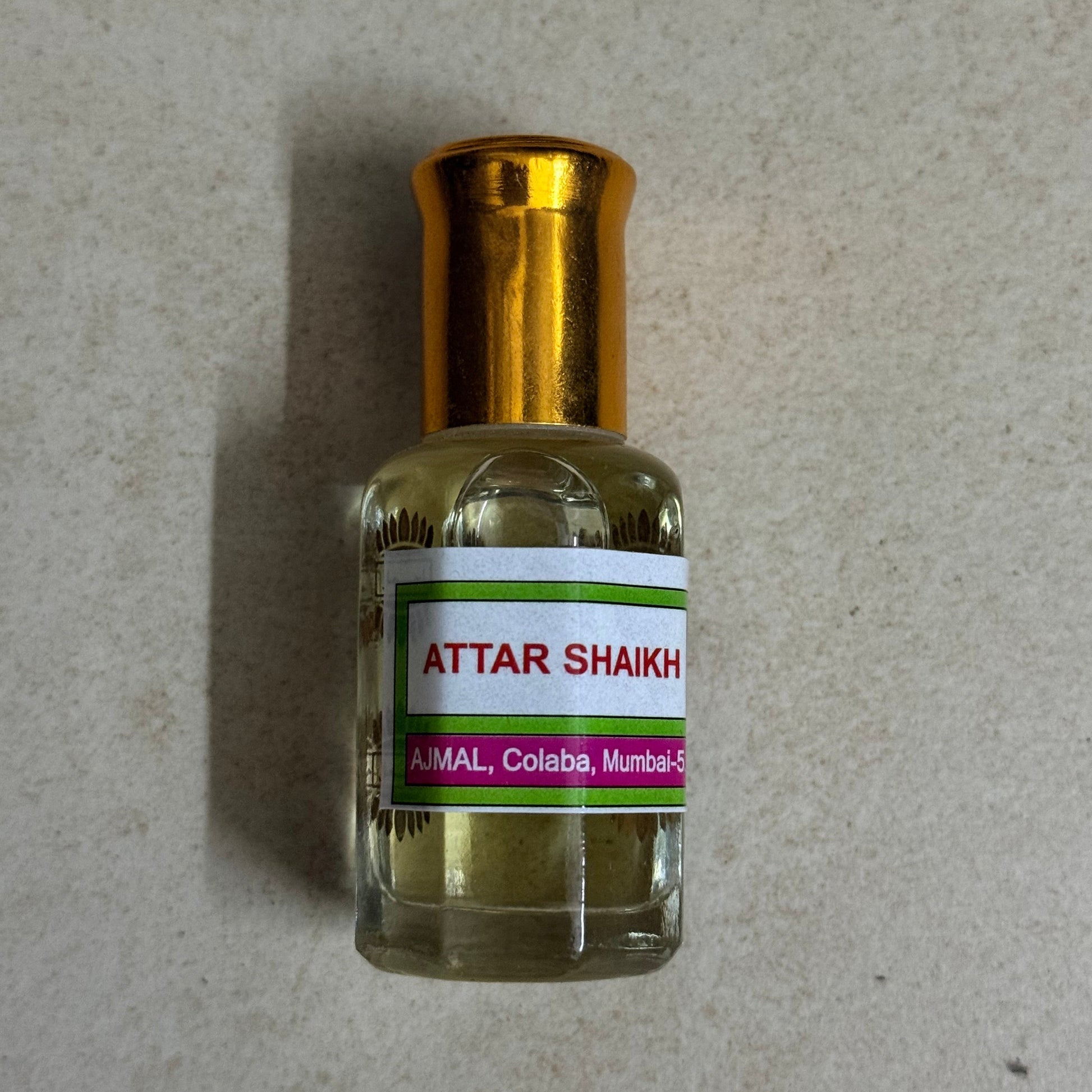 A small glass bottle of Attar Shaikh oil with a shiny gold cap, resting on a light gray textured surface.