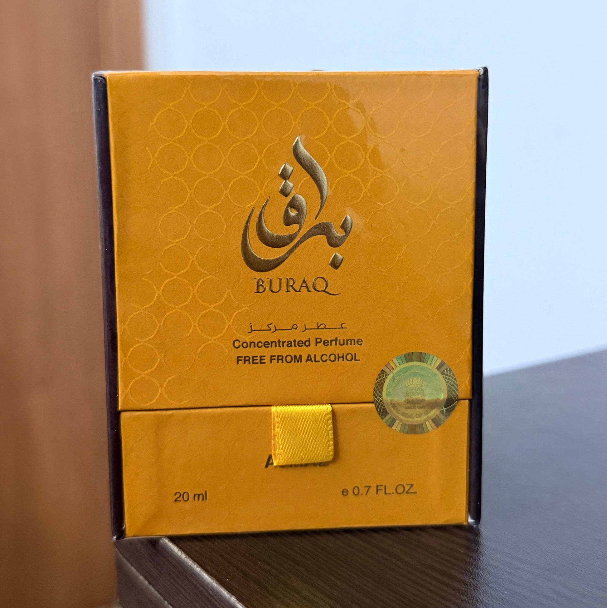 A rectangular Buraq perfume box in metallic gold and amber tones, standing on a dark wooden surface against a light blue wall background. The box features the Buraq brand name in English and Arabic calligraphy, along with 'Concentrated Perfume' and 'FREE FROM ALCOHOL'.