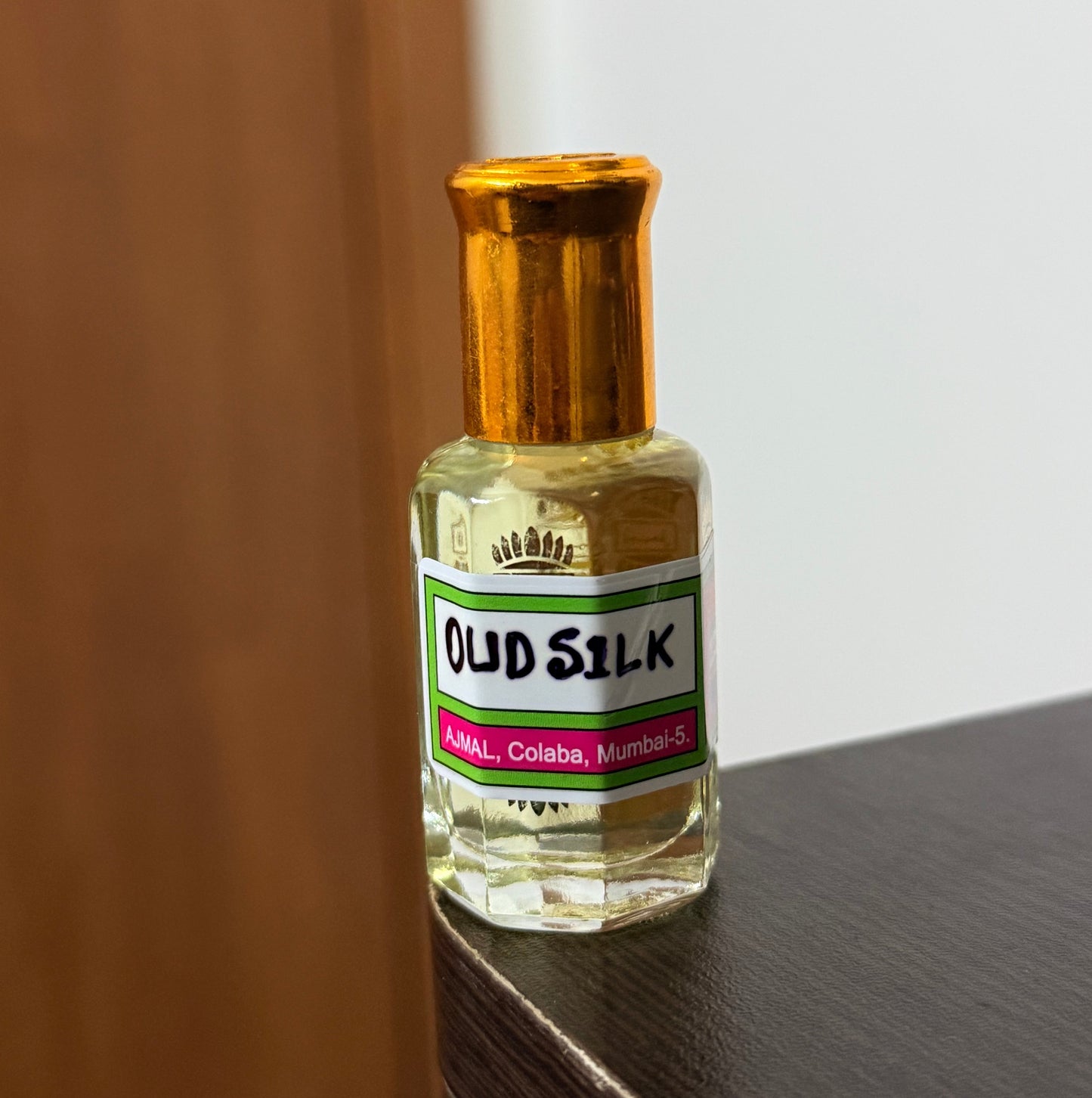 A small bottle of Old Silk oil with a gold cap and a white label, sitting on a dark wooden surface against a white wall.