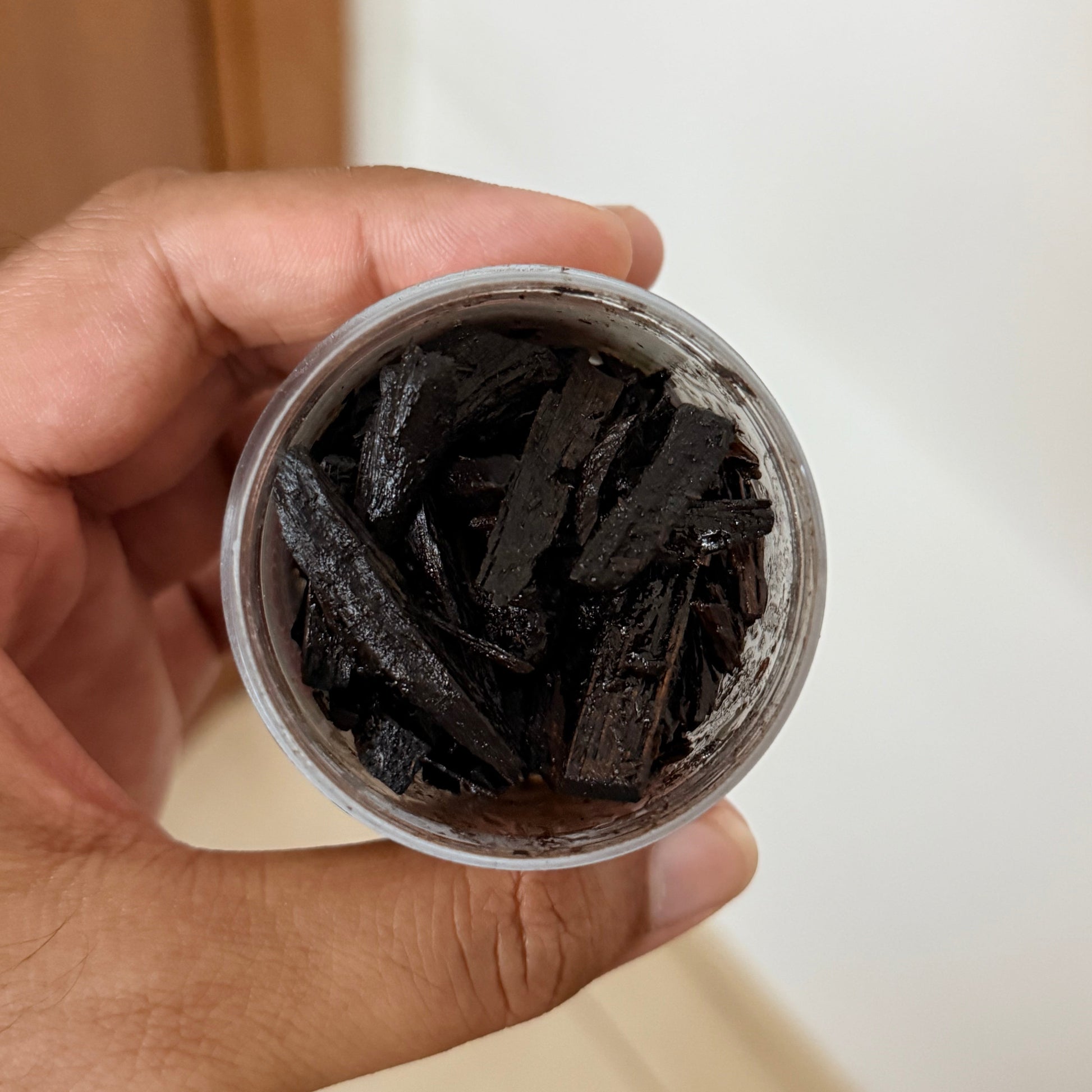 A hand holding a small clear plastic jar filled with dark brown to black shredded wood chips or bark pieces, photographed against a softly blurred indoor background.