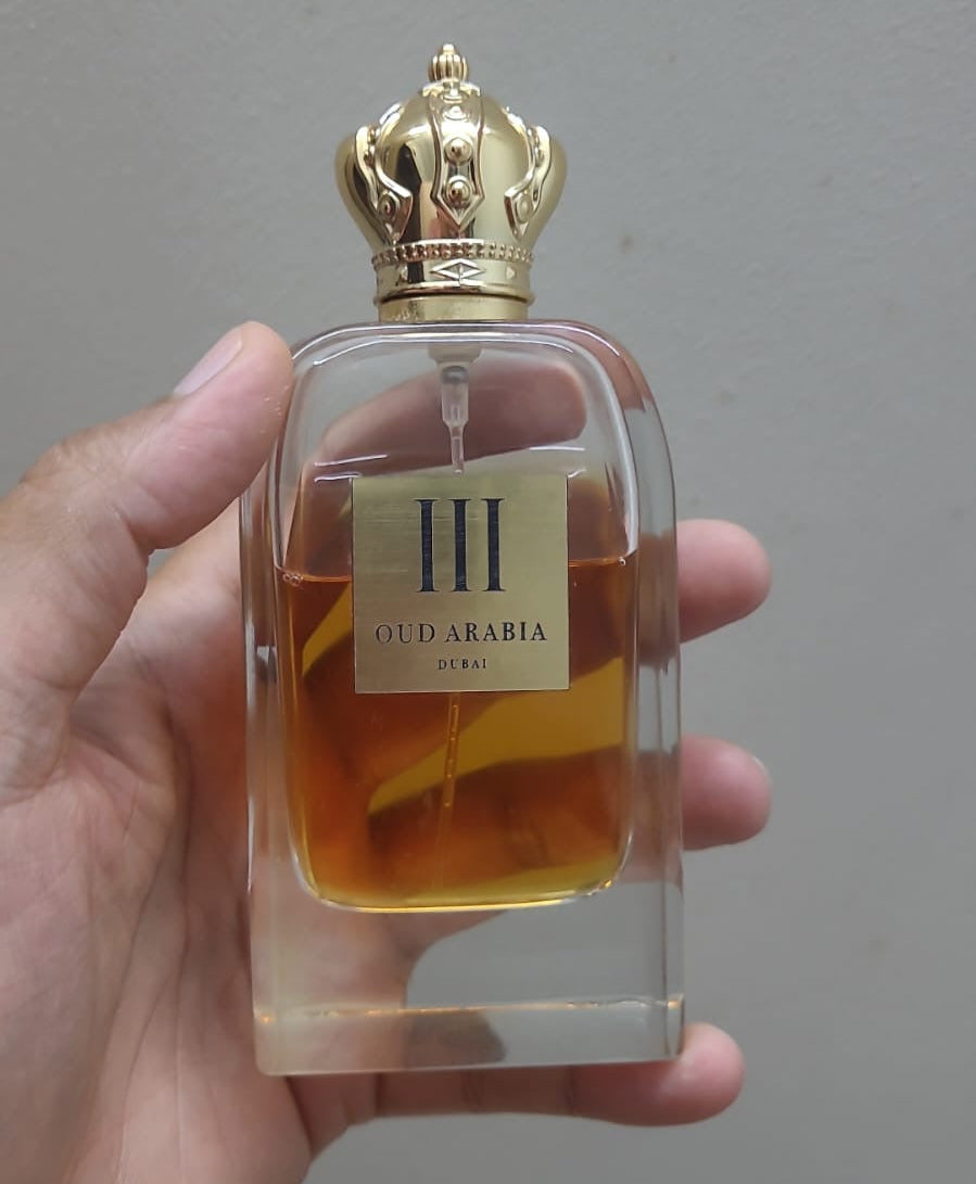 A hand holding a clear glass perfume bottle labeled 'III OUD ARABIA' with amber liquid inside and a gold crown cap, against a plain gray wall.