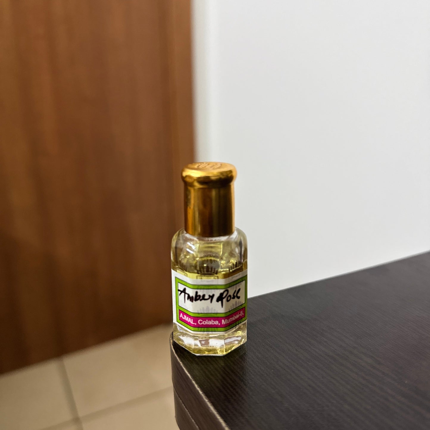 A small clear glass bottle with a gold cap and a white label reading 'Amber Rose' resting on the corner of a dark wooden table.