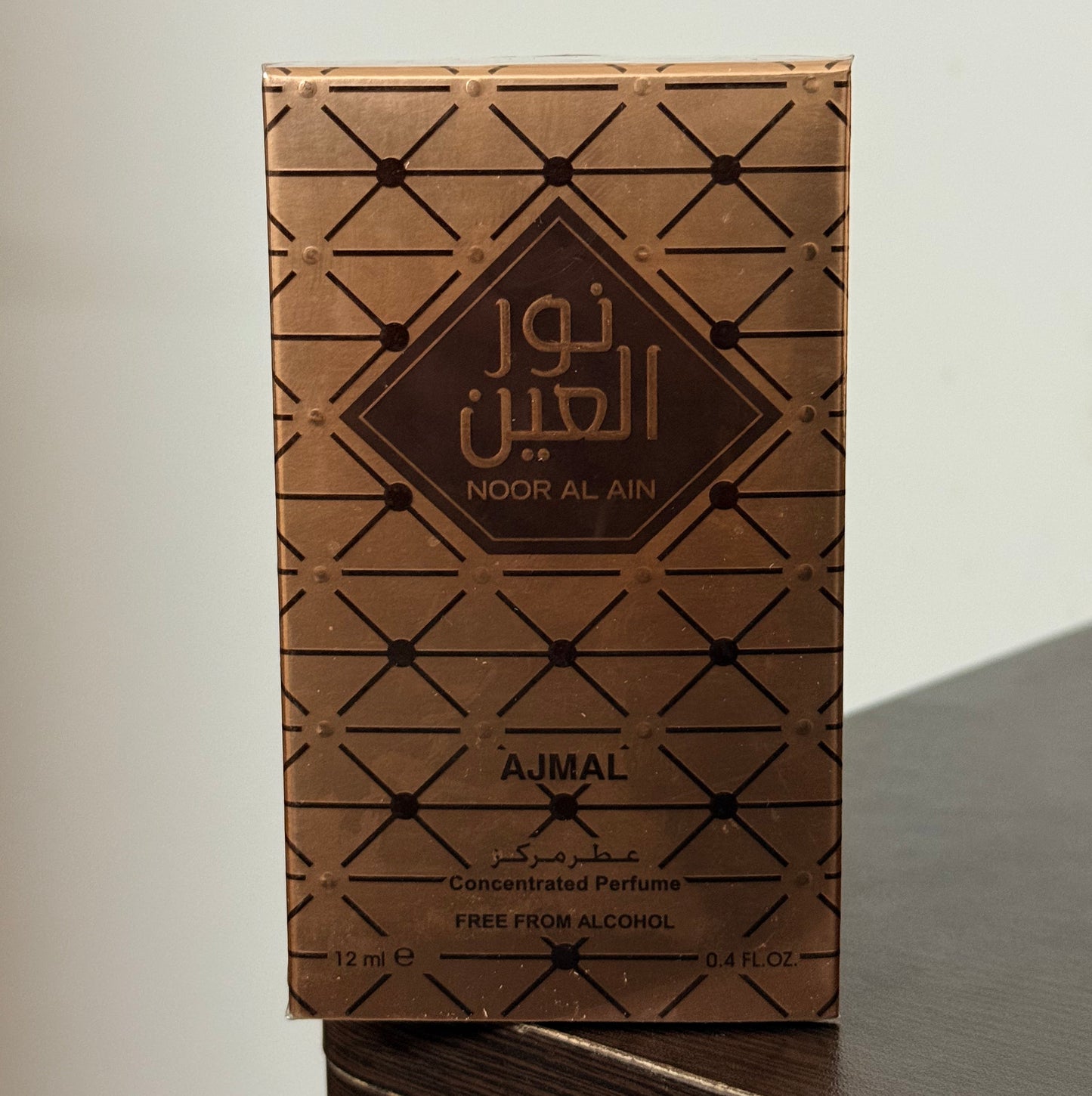 A rectangular perfume box for Noor Al Ain by Ajmal, featuring a brown and gold geometric pattern, resting on a dark wooden surface against a plain light background.