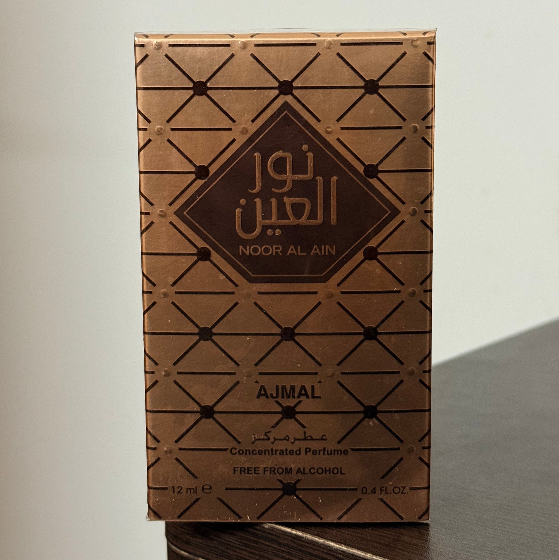 A rectangular perfume box for Noor Al Ain by Ajmal, featuring a brown and gold geometric pattern, resting on a dark wooden surface against a plain light background.