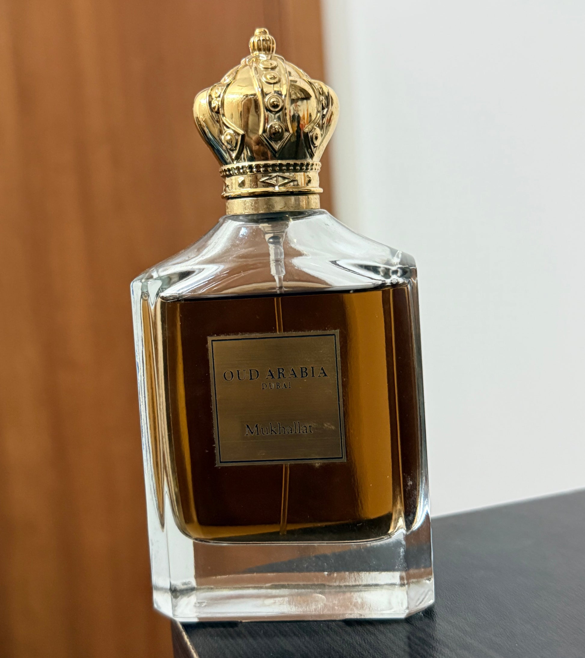 A bottle of Oud Arabia Mukhallat Parfum with a gold crown cap, set on a dark wooden surface against a background of a wooden door and a white wall.