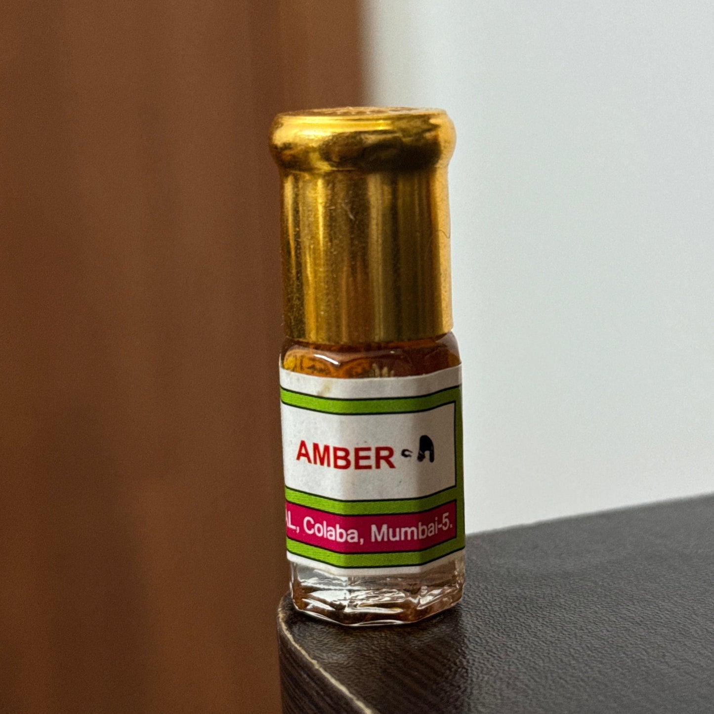 A close-up of a small amber glass bottle with a white, green, and red label reading 'AMBER-A' and 'Colaba, Mumbai', resting on a dark surface against a light background.