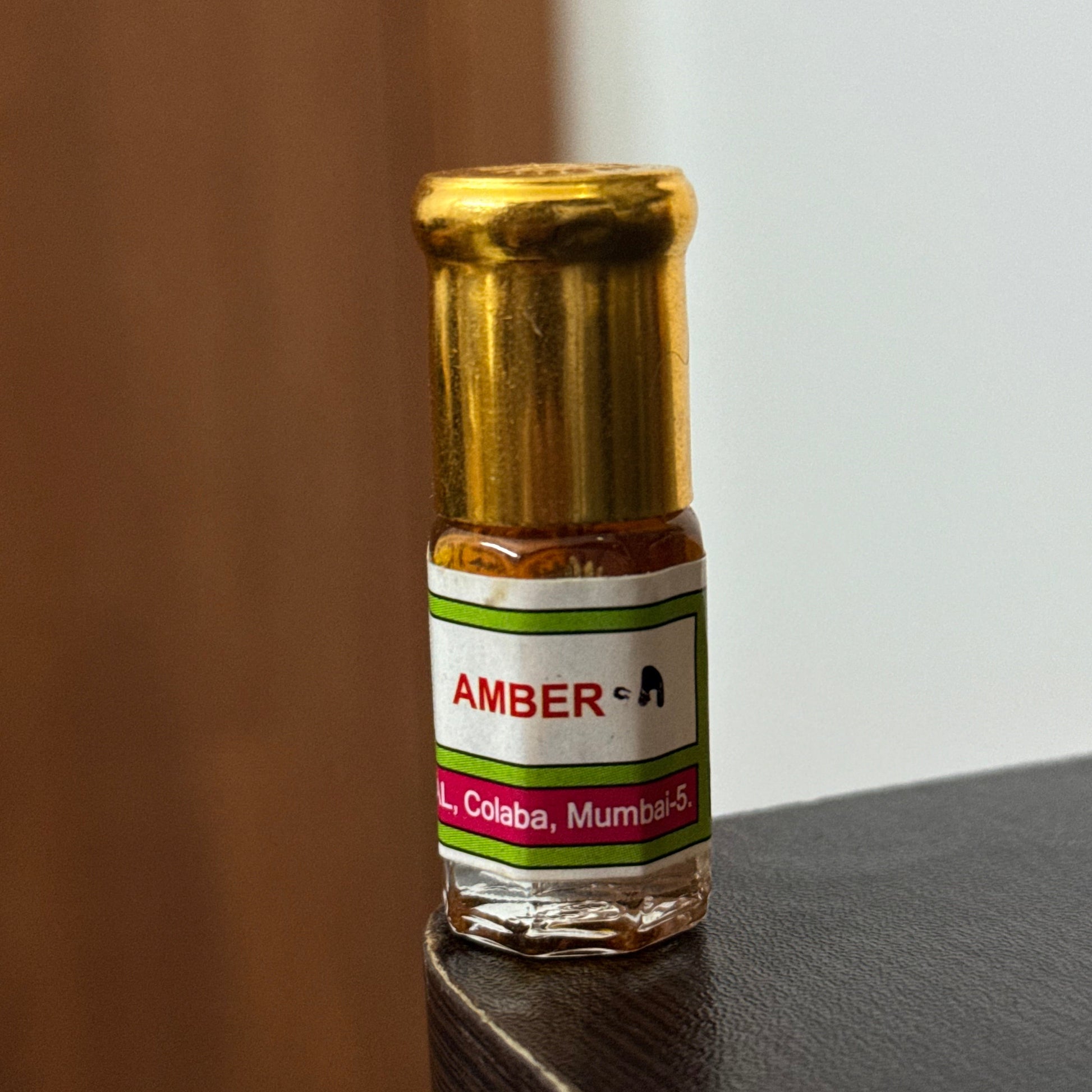 A close-up of a small amber glass bottle with a white, green, and red label reading 'AMBER-A' and 'Colaba, Mumbai', resting on a dark surface against a light background.