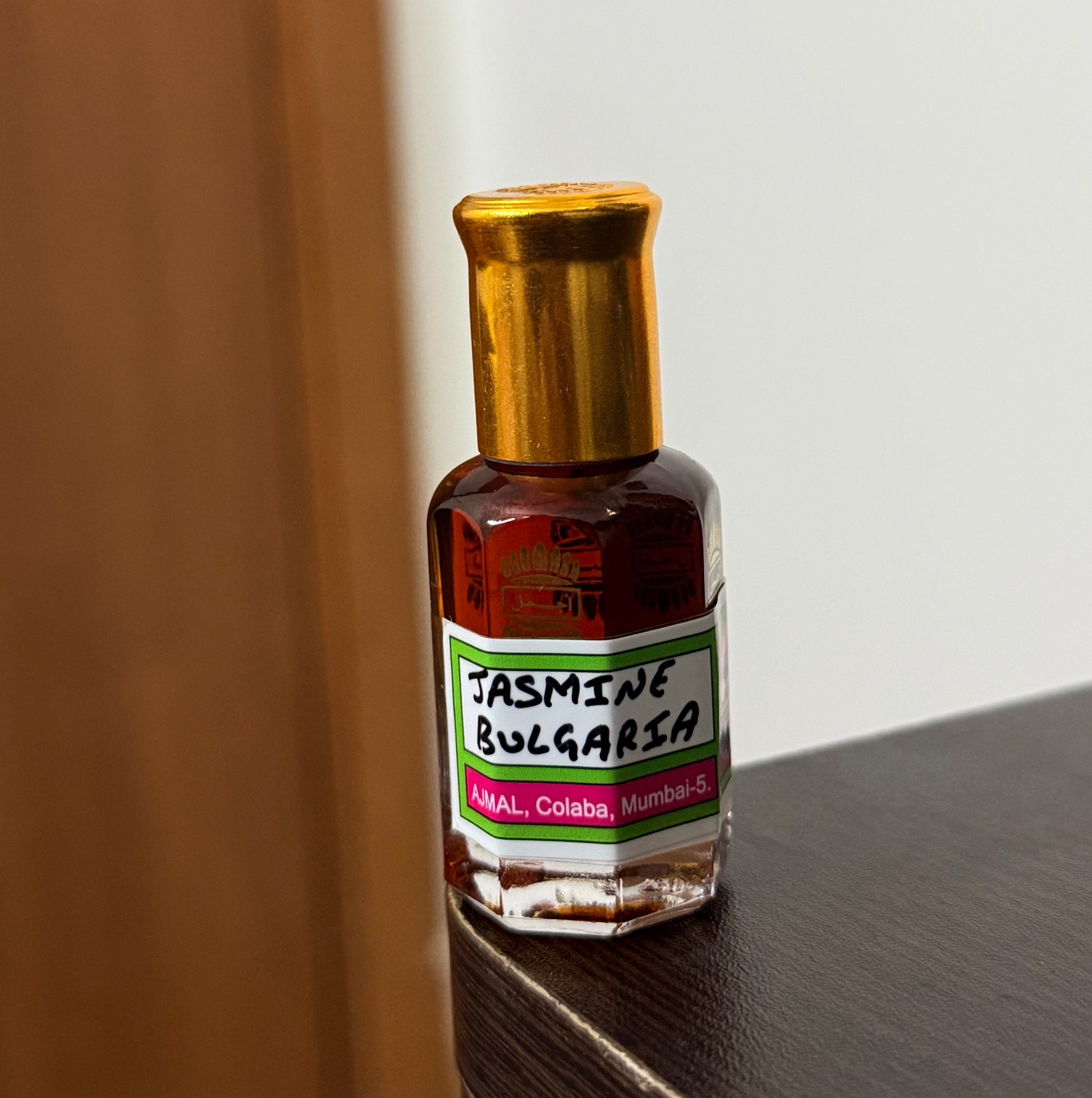 A small bottle of Jasmine Bulgaria essential oil with a gold cap and a white, green, and red label, resting on a wooden surface against a white wall.