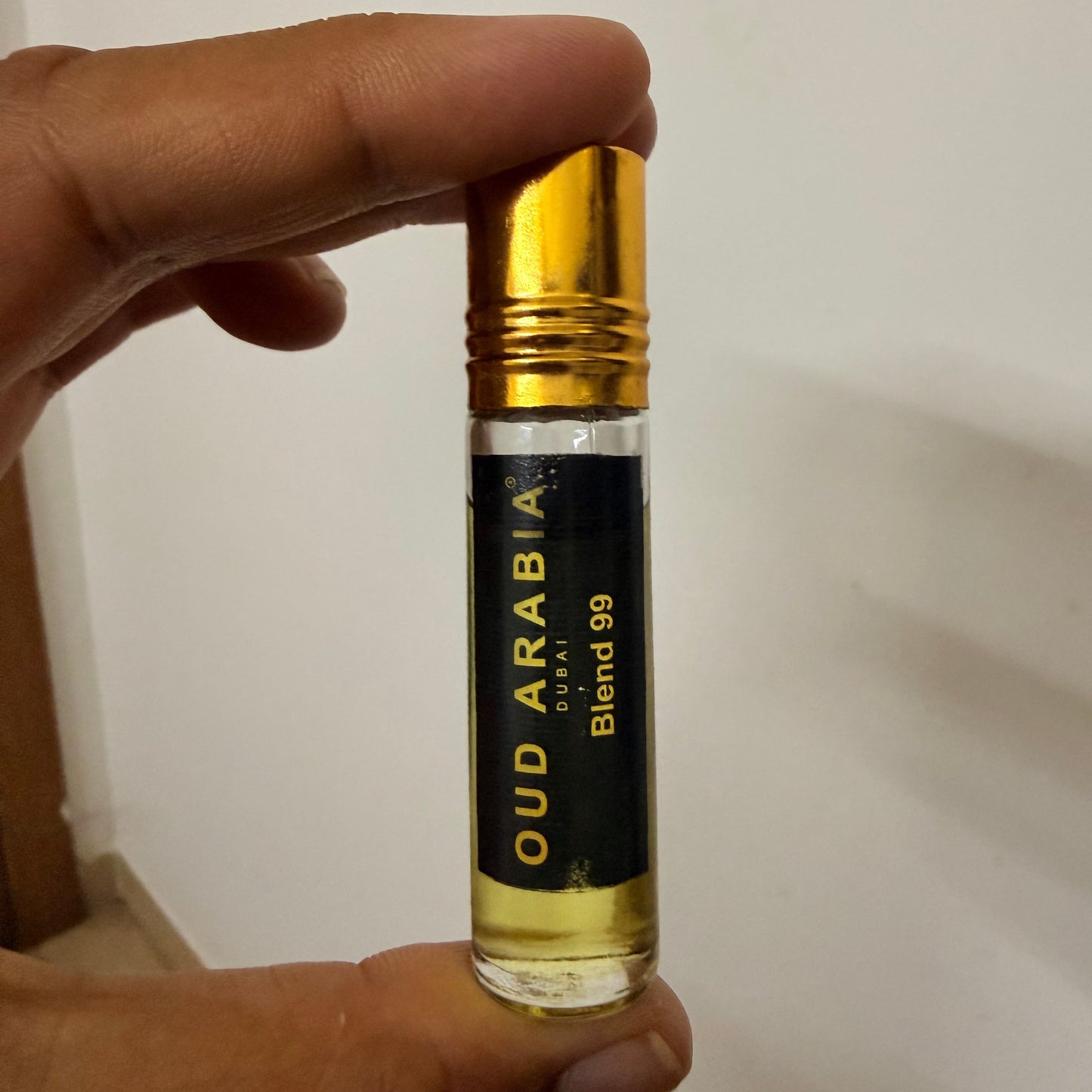 A hand holding a small cylindrical glass bottle of Oud Arabia Darwiq Blend 99 perfume oil against a plain off-white wall.
